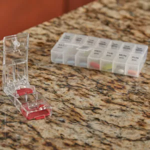 Angled view of opened red MAXSPLIT™ Safety Pill Splitter with Puncher on a marble top next to a weekly pill organizer | Maxpert Medical